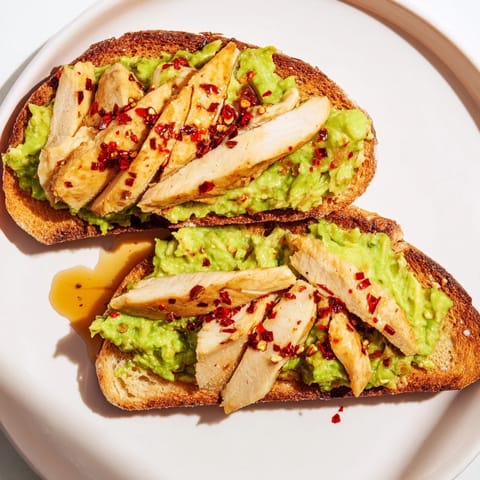 Golden grilled chicken rests on vibrant green avocado mash spread over crusty toast, sprinkled with chili flakes and drizzled with honey.