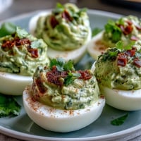 Guacamole Stuffed Deviled Eggs with creamy avocado filling, garnished with smoked paprika and fresh cilantro on a white platter.